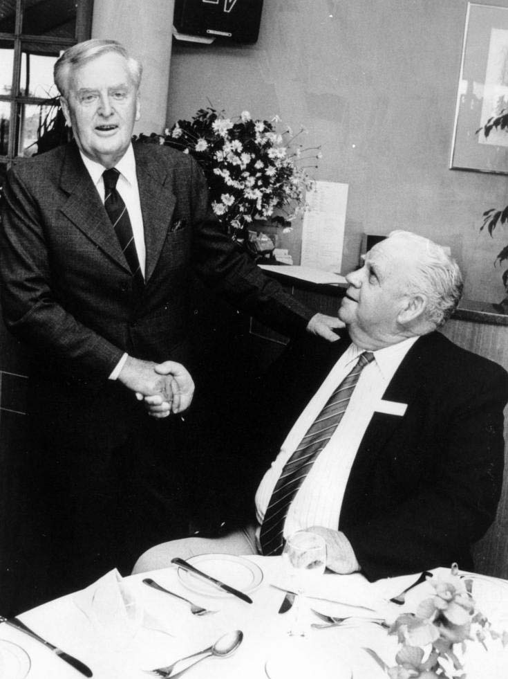 B&W photo of Sir Joh Bjelke-Petersen shakes hands with a seated Russell Hinze on October 11, 1989.