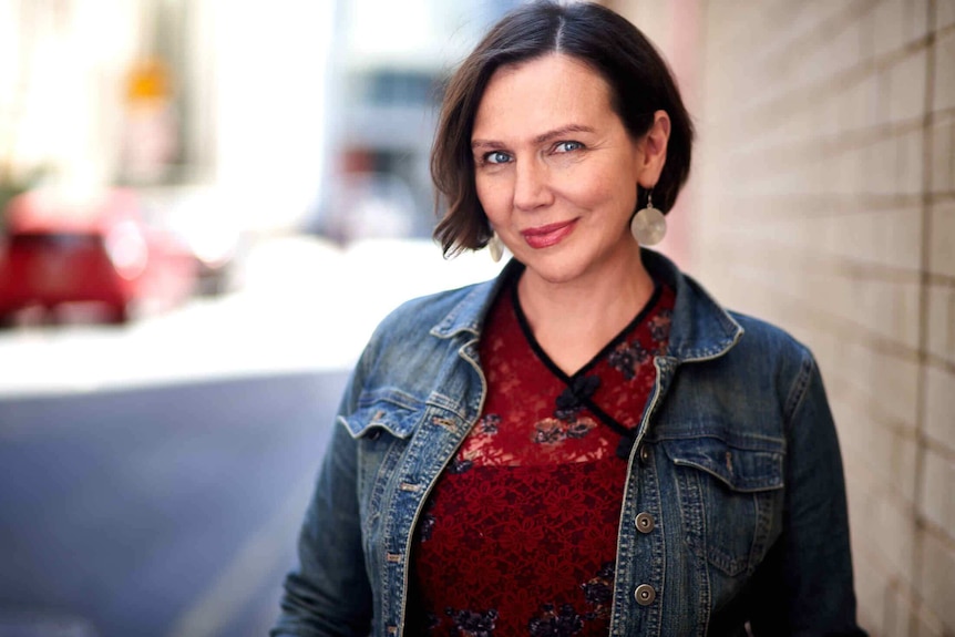 Before a blurred, bright streetscape, a woman with short hair, drop-earrings, denim jacket and red print jumper smiles.
