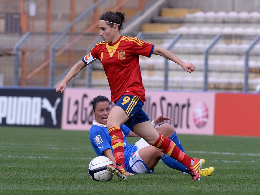A soccer player wearing red, yellow and blue kicks the ball during a game while an opposition player is on the ground