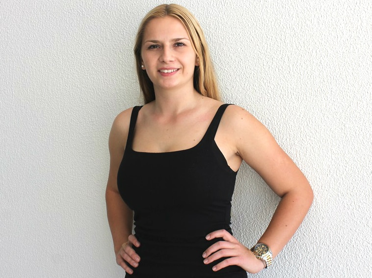 Woman in her early 20s stands looking at the camera, with long blonde hair, smiling.