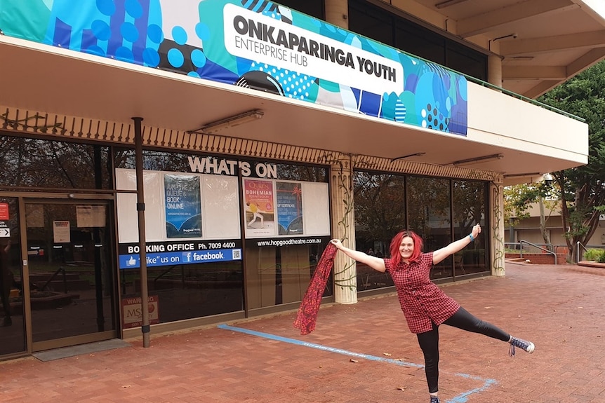 A young woman with red dyed hair throws her arms up and smiles outside a youth hub building.