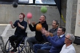 Seven people in wheelchairs in a cement room throwing coloured basketballs in the air