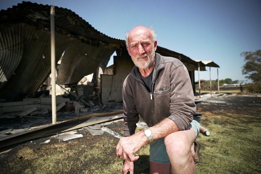 A man with white hair kneels in front of his destroyed house.