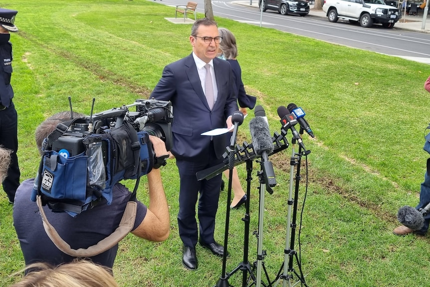 A man in a suit speaking to microphones with a TV camera in a park