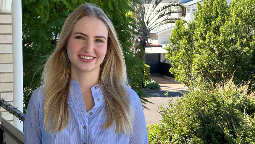 Naomi Lewis smiles while standing on a balcony in front of a tree-lined street. She has long blonde hair and wear a blue shirt.