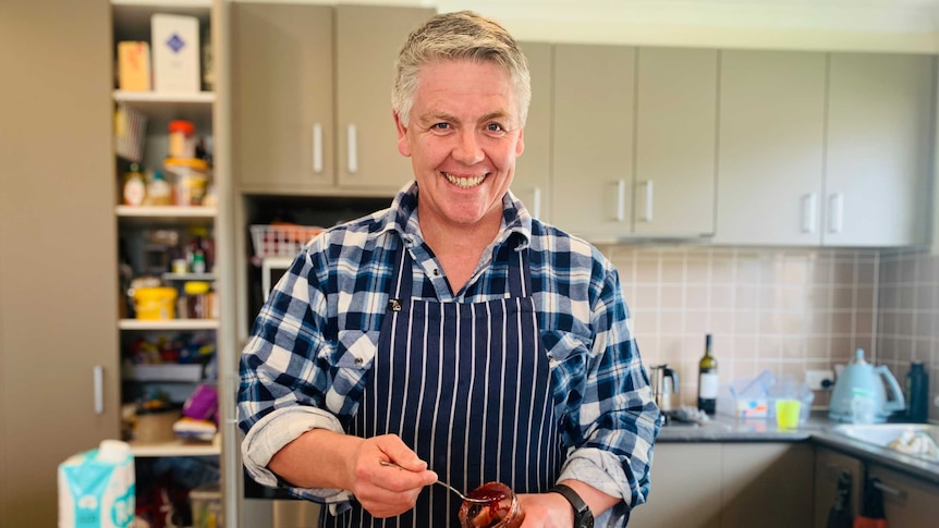 Male cook stands in his kitchen holding a spoon to spread homemade strawberry jam on his freshly baked bread.