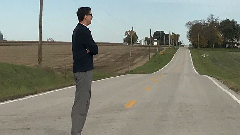 James Comey stands on a road looking out across fields in Iowa.