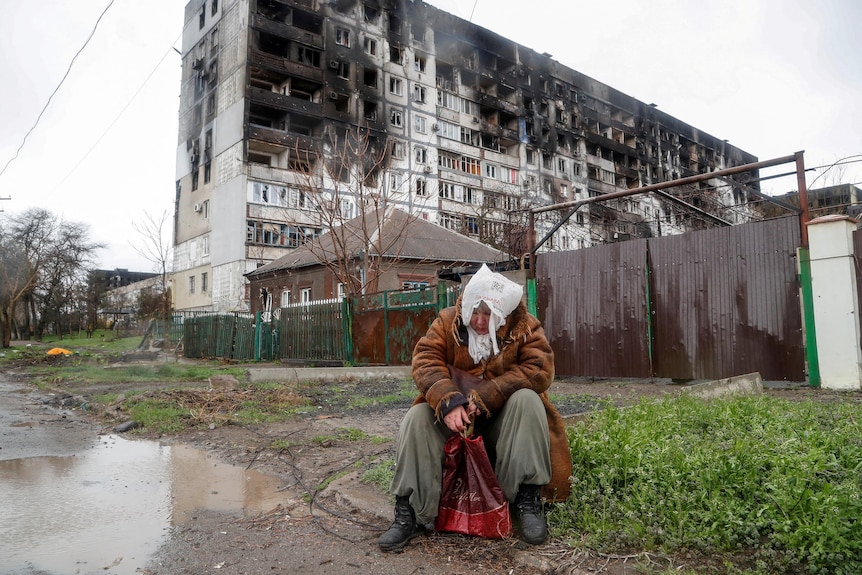 Женщина сидит перед сильно поврежденным многоквартирным домом