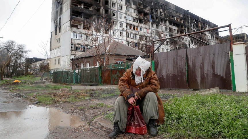 A woman sits in front of a heavily damaged residential building