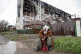 A woman sits in front of a heavily damaged residential building