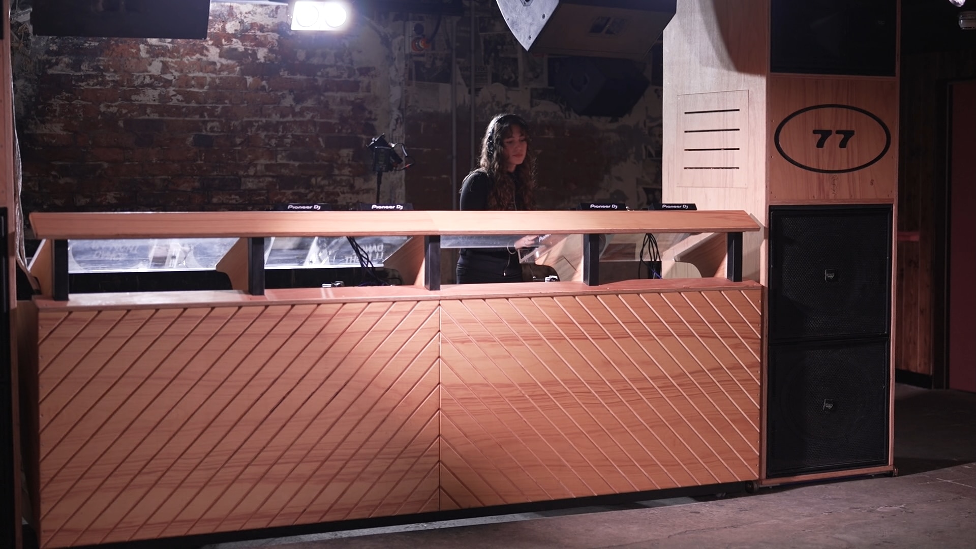 DJ decks across a wooden table panel, with a girl in headphones behind them.