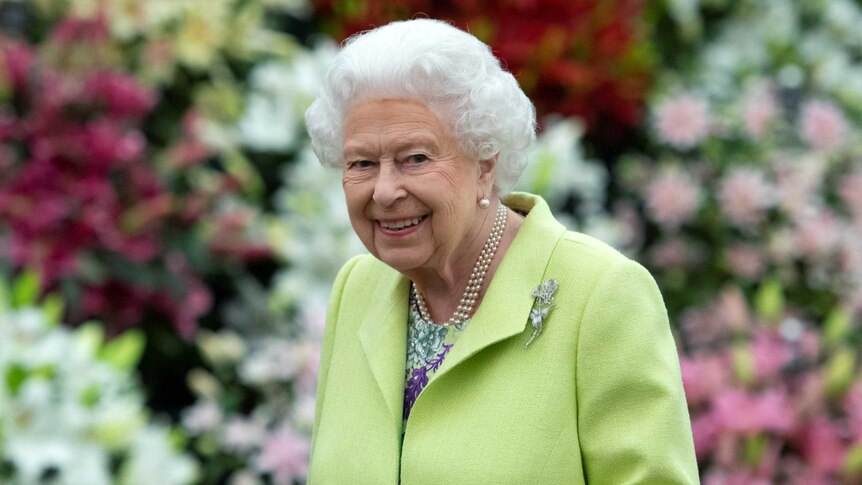 Queen Elizabeth II smiles at the camera in a floral garden, wearing a green coat and gloves. 