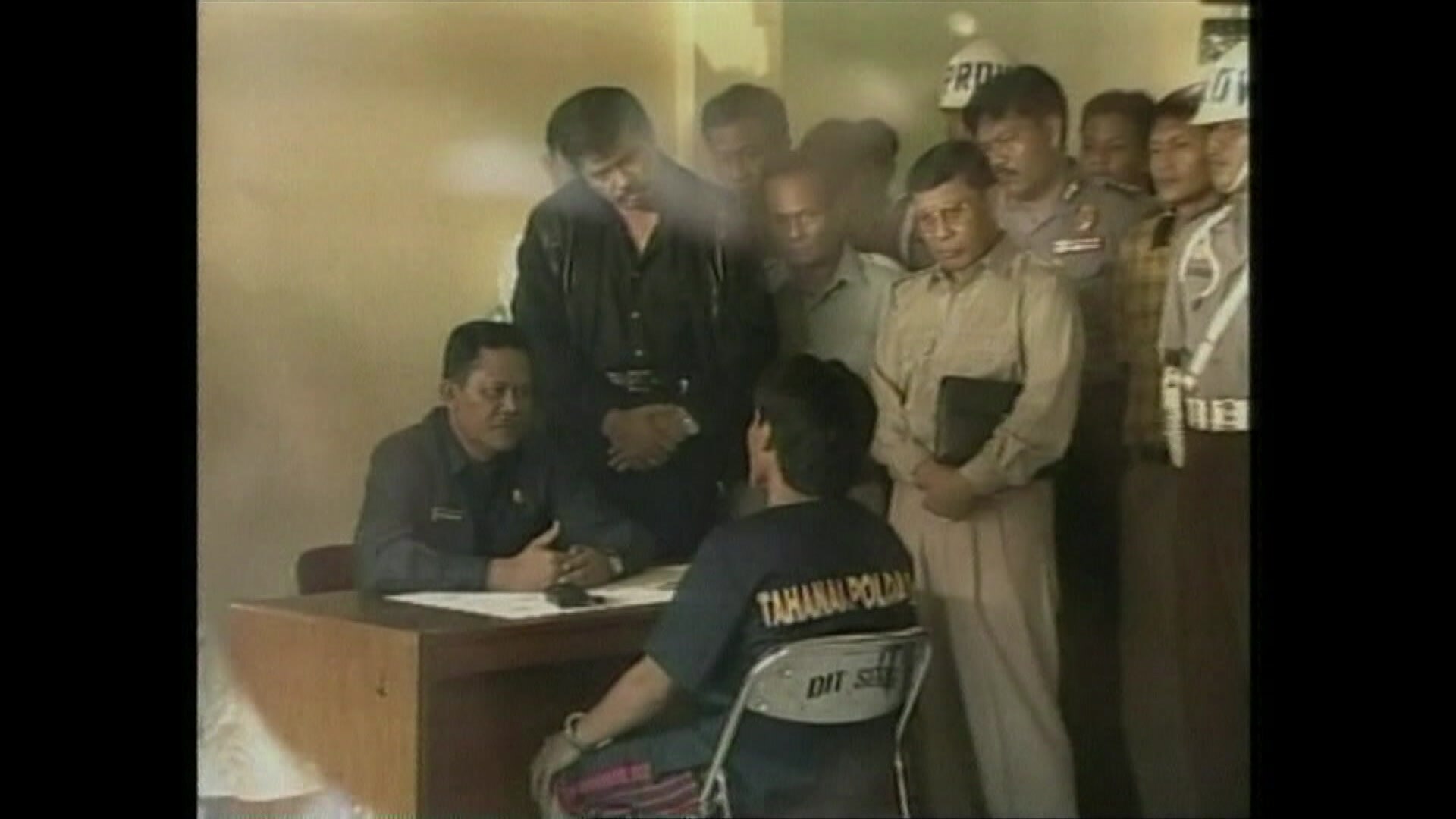 Two men sit facing each other at a small desk, one handcuffed, as a crowd of men lean over them.
