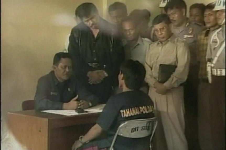 Two men sit facing each other at a small desk, one handcuffed, as a crowd of men lean over them.