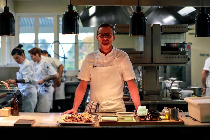 Chef wearing white uniform stands at bench beneath heat lamps with large kitchen and chefs behind him