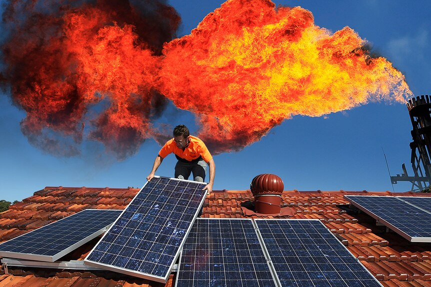 A worker installing rooftop solar panels with a industrial chimney venting a big flame behind.