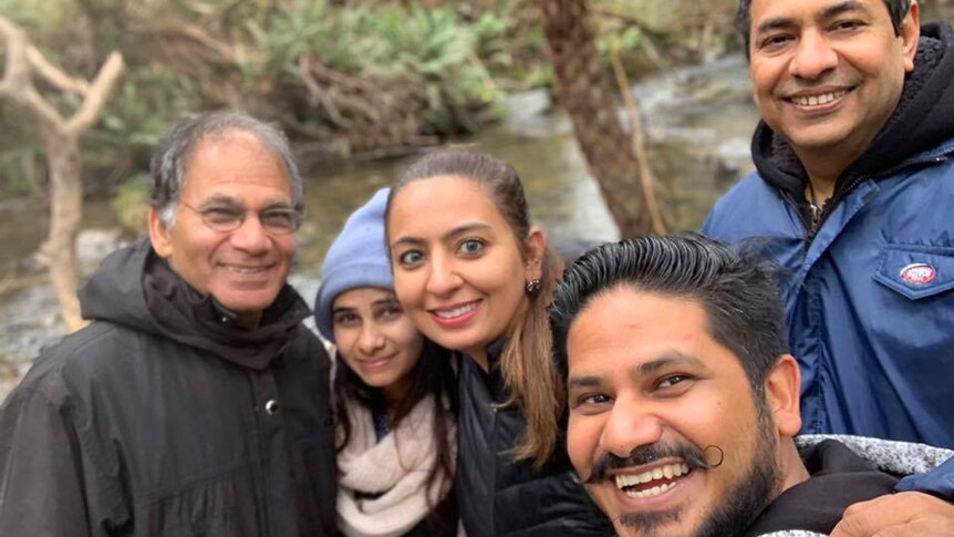 The Gupta family — two women and three men — poses for a photo together. They are smiling by a riverbank.