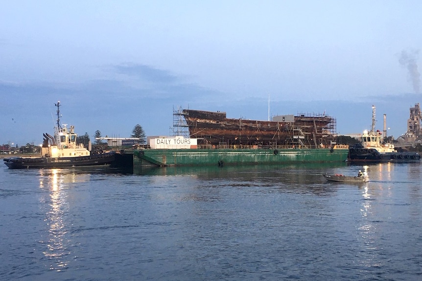 The hull of an aging ship sits on a barge and is pushed by a tug boat.
