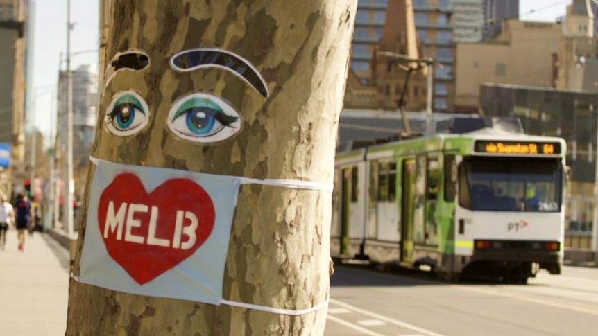 A blue mask with a red heart and 'MELB' printed on it is attached to a tree on the side of the footpath at Southbank, Melbourne.