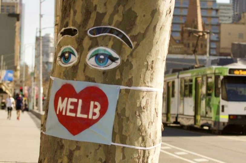 A blue mask with a red heart and 'MELB' printed on it is attached to a tree on the side of the footpath at Southbank, Melbourne.