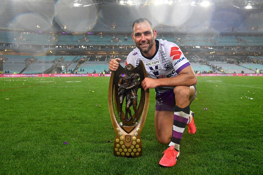 A smiling NRL player kneels on ground holding the premiership trophy in the rain at the grand final.