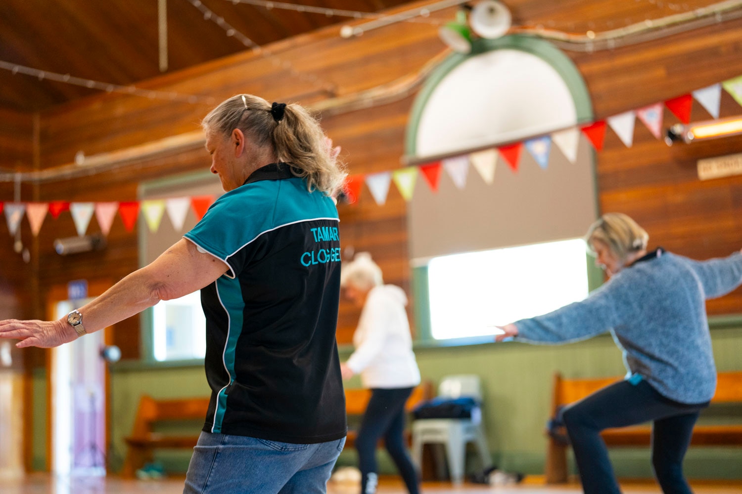 women dance in tap shoes in black and teal shirts in a wooden town hall