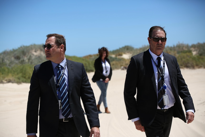 Detectives in suits and sunglasses walk along a sandy coastal area