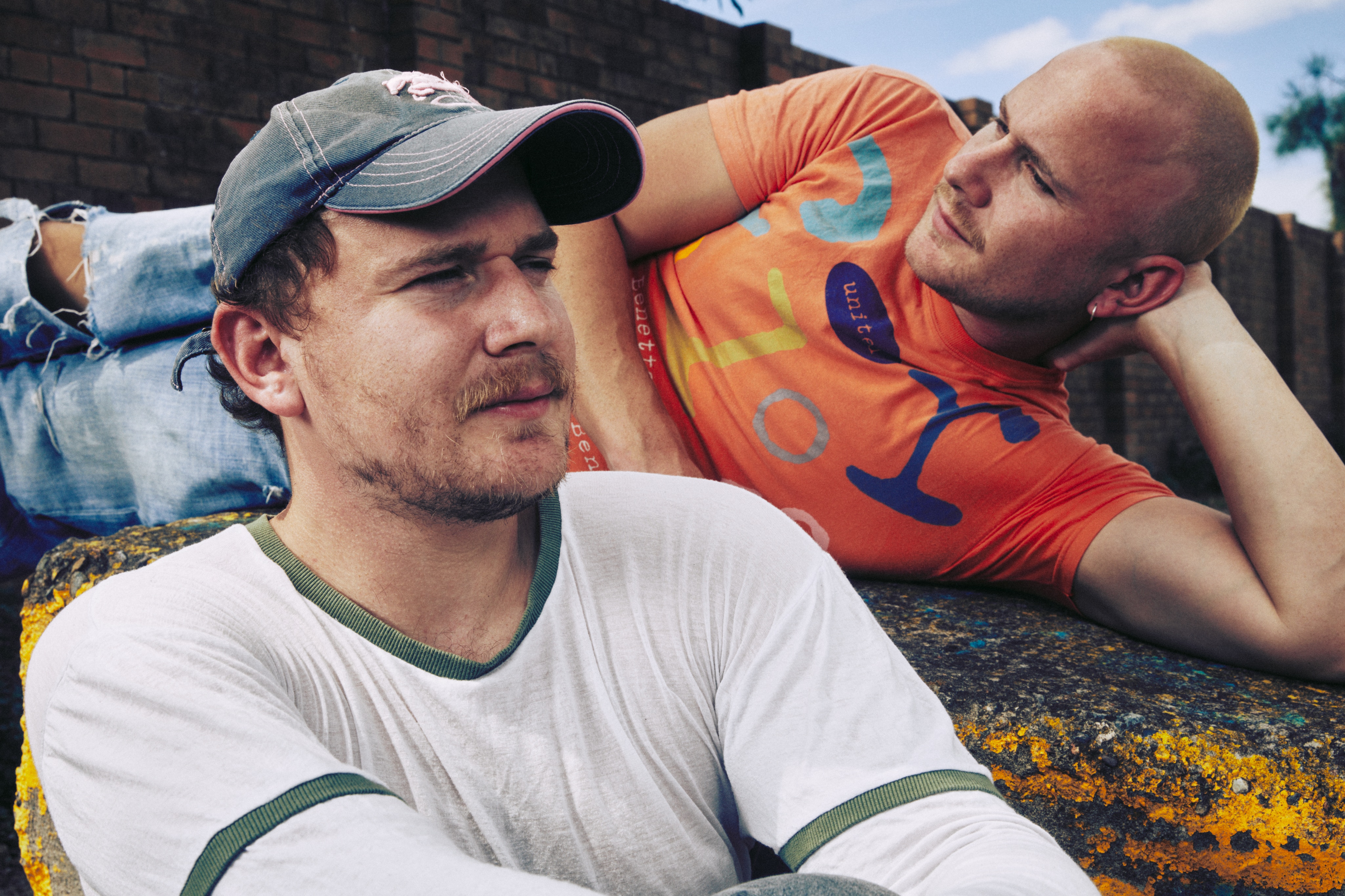 Close up photo of twin men, one in a grey cap looking to the right and one lying down in an orange shirt looking to the left