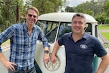 Two men stand in front of a blue and white coloured Kombi.