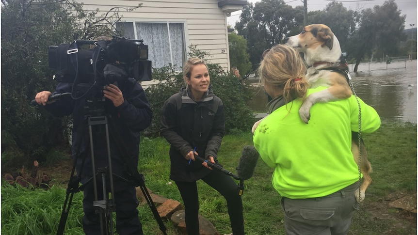 Journalist holding boom microphone interviewing a woman holding a dog with floodwaters in background.