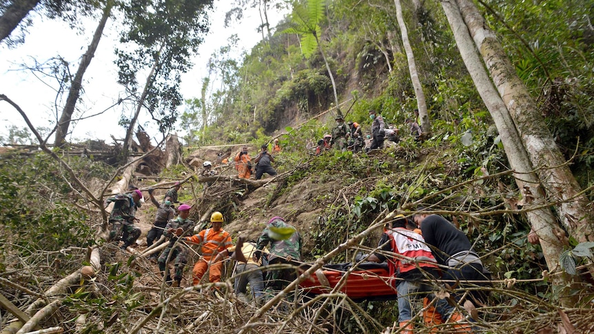 A narrow trail zig-zags up a steep hill in rainforest with rescue workers in orange carrying a body on a spinal board.