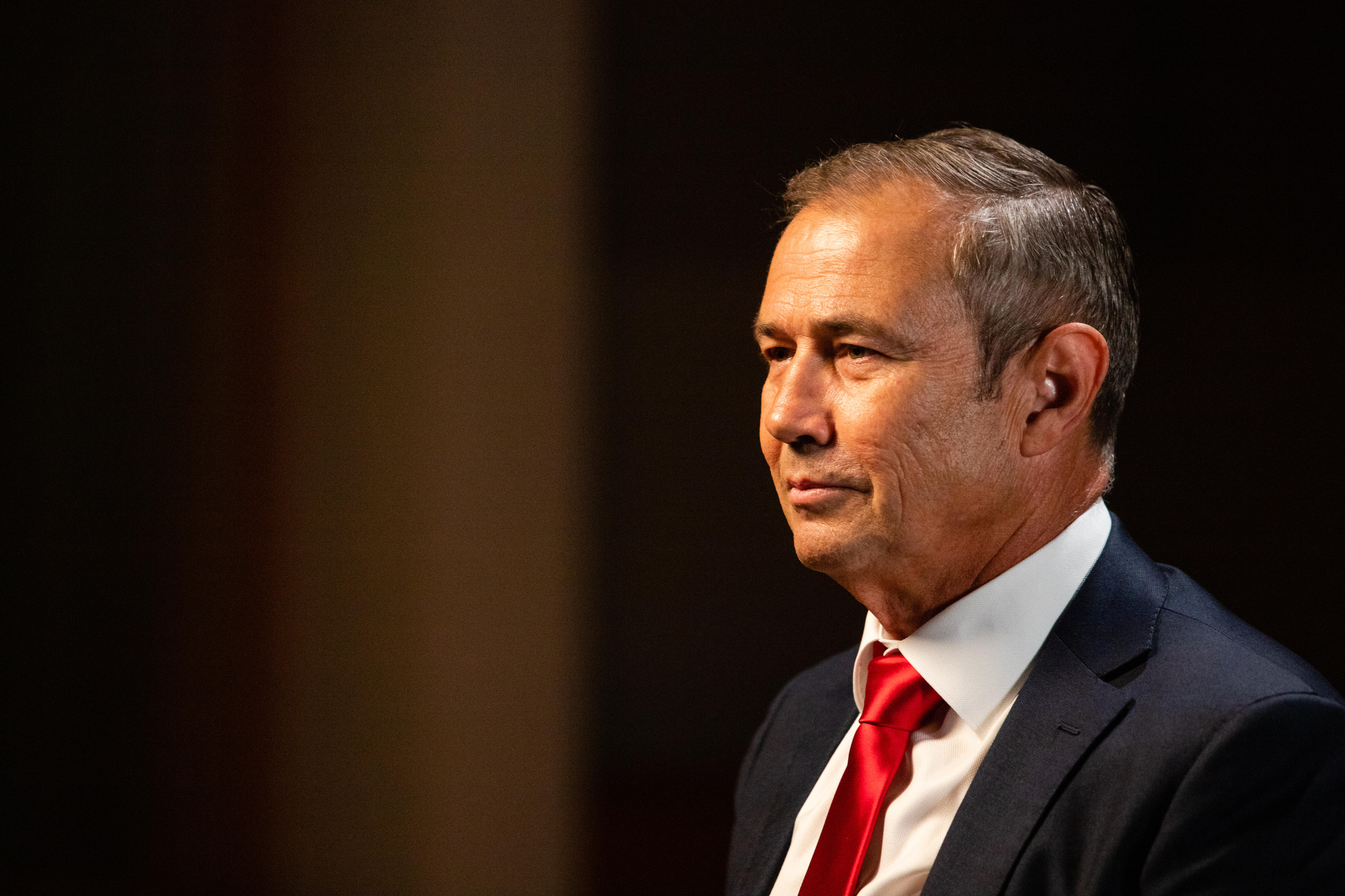 A man in a dark suit with a red tie sits in a darkened room.