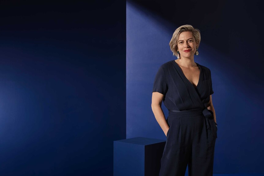 Portrait of a striking middle aged woman with blonde hair, red lipstick, wearing a navy jumpsuit against a blue backdrop.