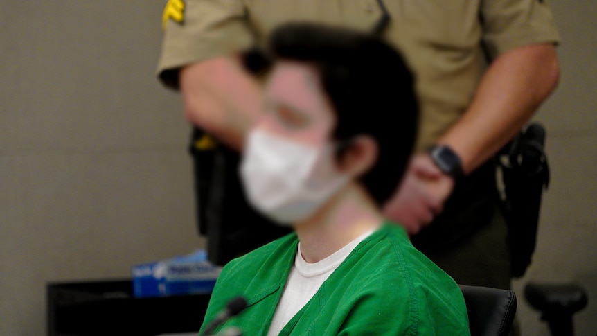 The blurred face on a man in a green prison outfit. A sheriff stands behind him.