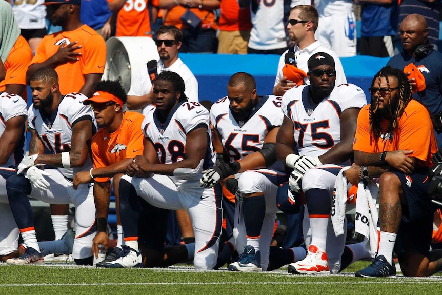 Six footballers kneel on the sideline during a pre-game national anthem.