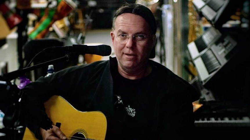 A tired-looking Scott Burford plays his guitar while sitting on a stool inside the Engadine music shop.