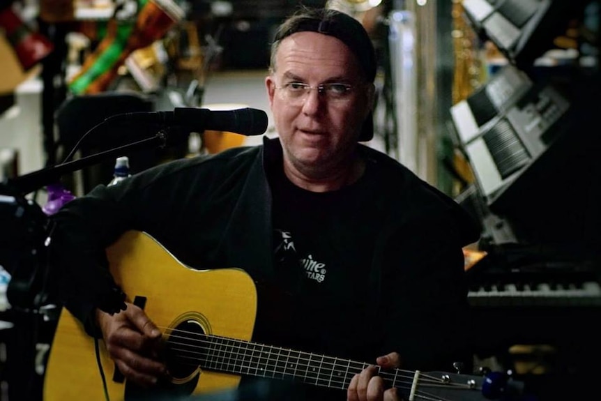 A tired-looking Scott Burford plays his guitar while sitting on a stool inside the Engadine music shop.