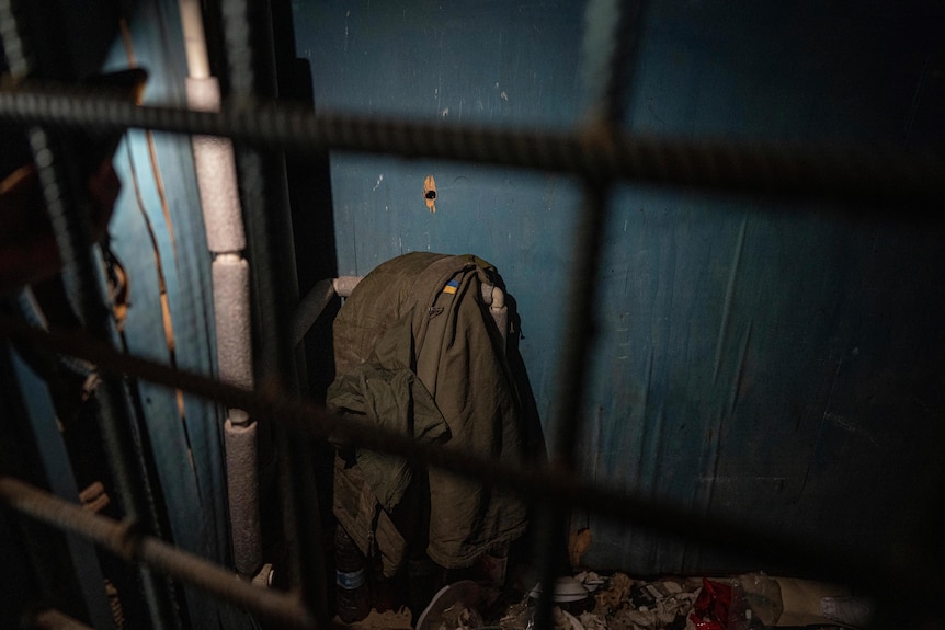 An army jacket bearing a patch with Ukraine's flag is seen hanging over a pipe in a blue-walled cell room.