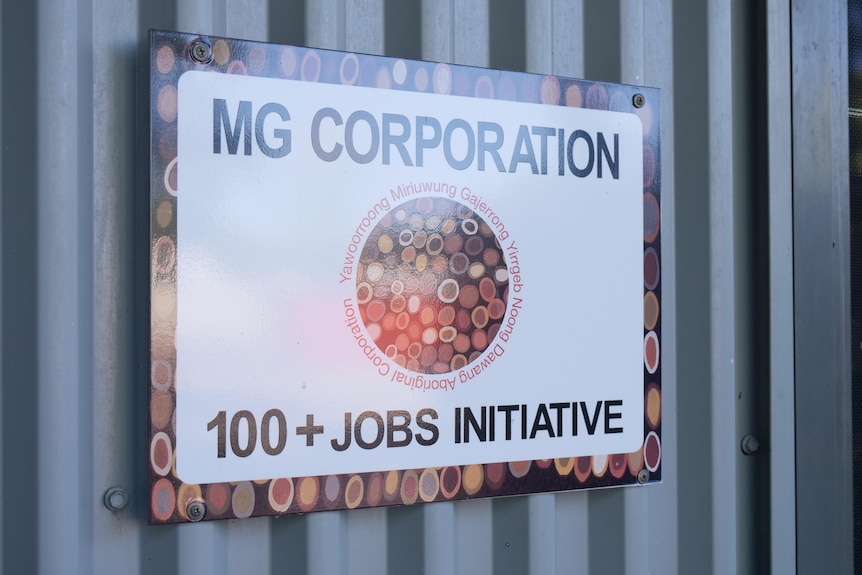 Sign on side of shed, saying 'MG Corporation' at the top and '100+ Jobs program' below an Aboriginal artwork