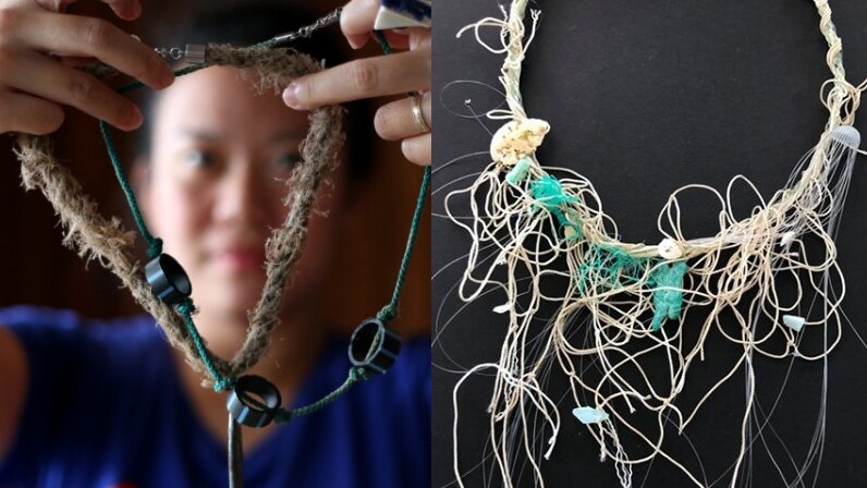 A woman holds a necklace made of rope, sinkers and plastic piping, and a necklace made of string and fishing line on a table.