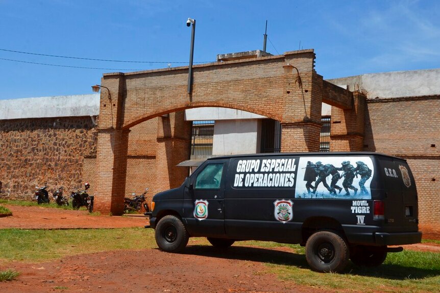 A large police black van with a photo of gunmen on its side is parked out of the front of an orange brick prison on a clear day.