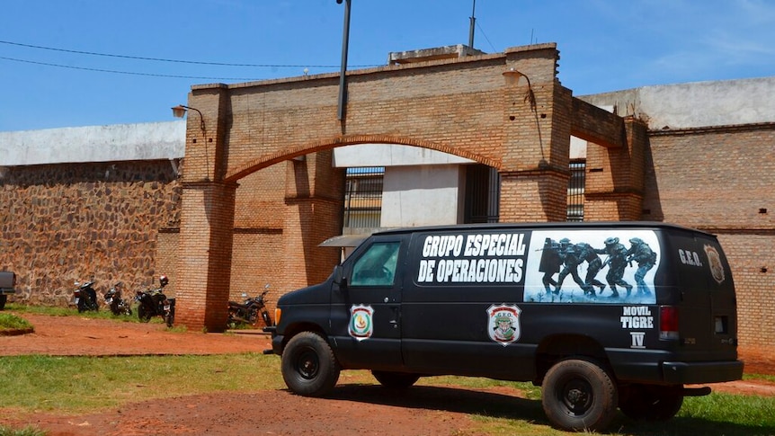 A large police black van with a photo of gunmen on its side is parked out of the front of an orange brick prison on a clear day.
