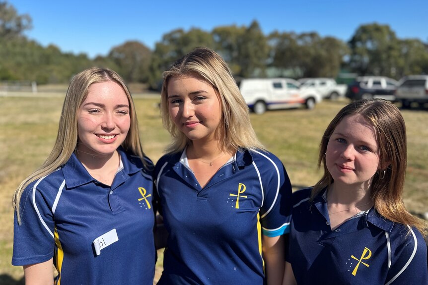 Three high school students in school uniform face the camera with a carpark in the background. 