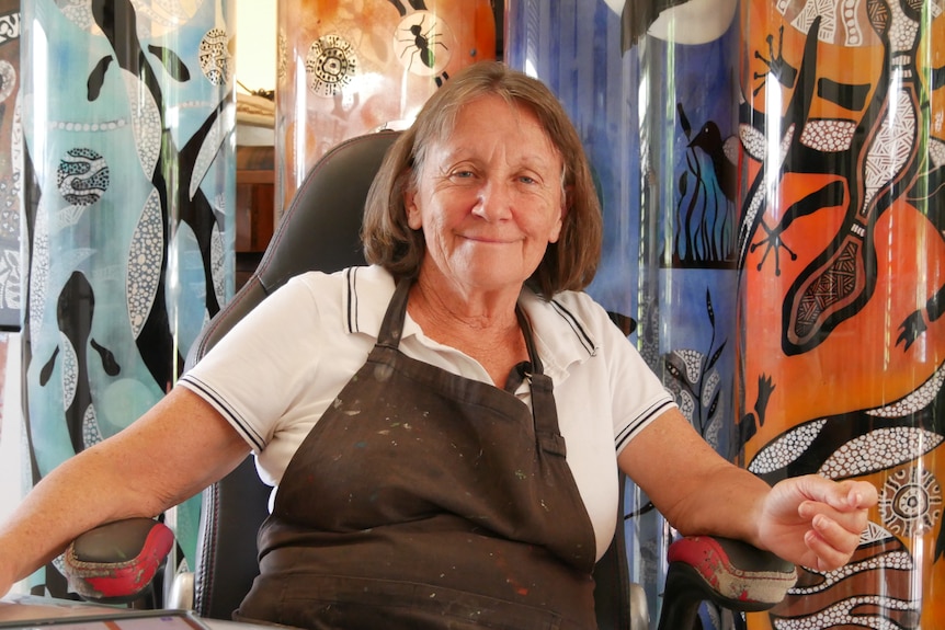 A woman with short light brown hair sits in front of sculptures featuring brightly coloured Indigenous artwork