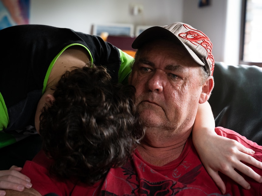 A crying man in cap is hugged by a boy whose face is obscured.