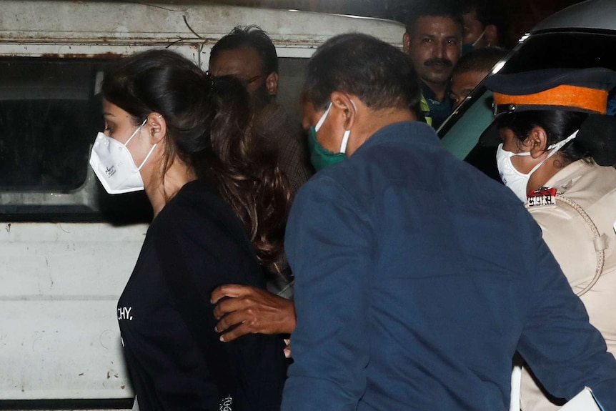 A Bollywood actor being being guided by a police official. She wears a mask and her hair in a ponytail.
