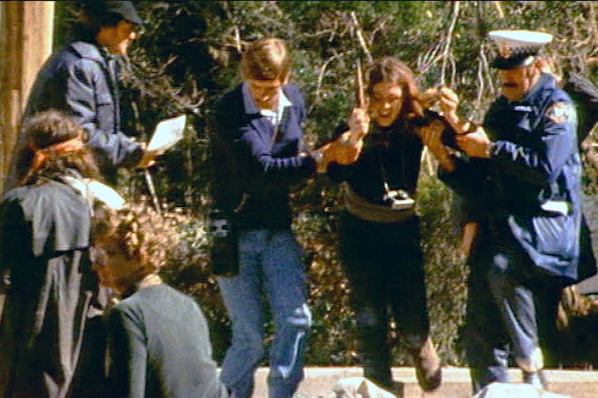 A woman in a black shirt is lead off by the arms by two police officers.
