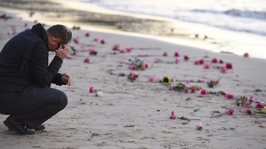 A man crouches by the waters' edge holding his head in his hands.