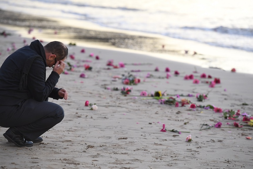 A man crouches by the waters' edge holding his head in his hands.