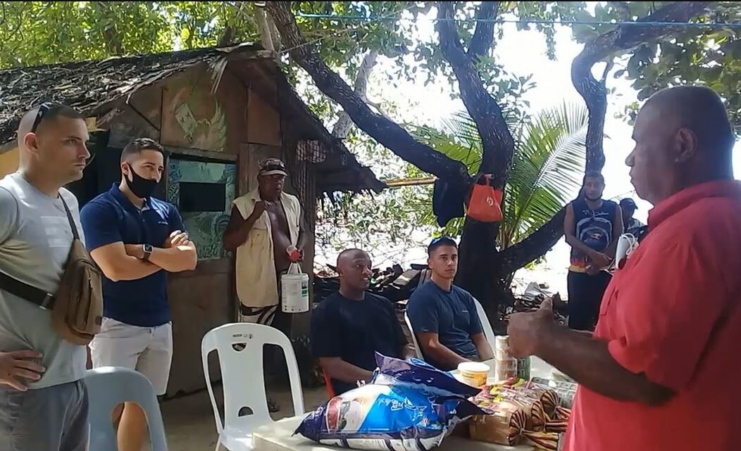 An older man speaks to two seated men and three standing men, gathered around a plastic table outside in shade.